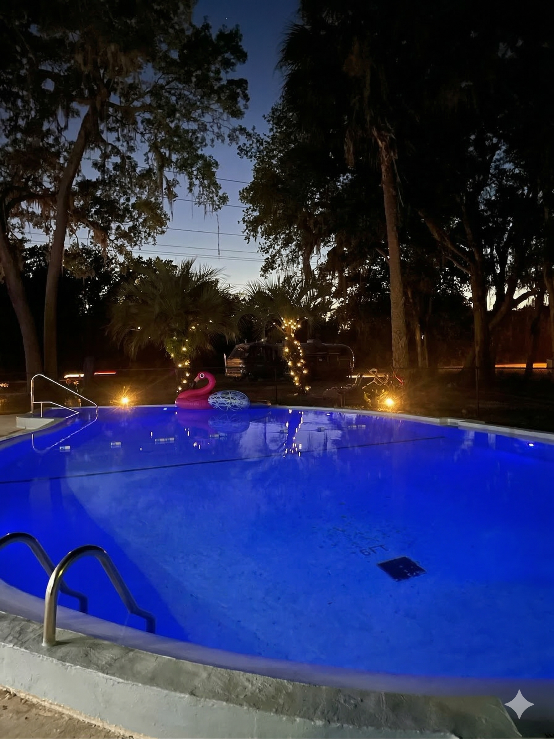 Blue-lit outdoor swimming pool at dusk with a pink flamingo float, surrounded by trees and string lights in the background.
